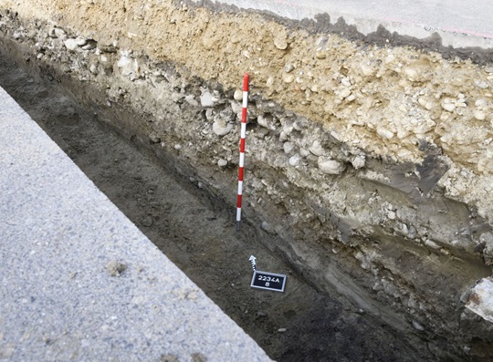 Im untersten Bereich des Profils im Leitungsgraben steht ein dunkles Lehmband an – ein ehemaliger Lehmestrich – und rechts der Fototafel ein dunkles Band, das einen geschnittenen Balkengraben darstellt.